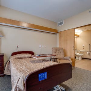 Newly remodeled nursing home facility room with medical bed and dresser to one side with rocking chair to other side and sliding door leading to bathroom behind.