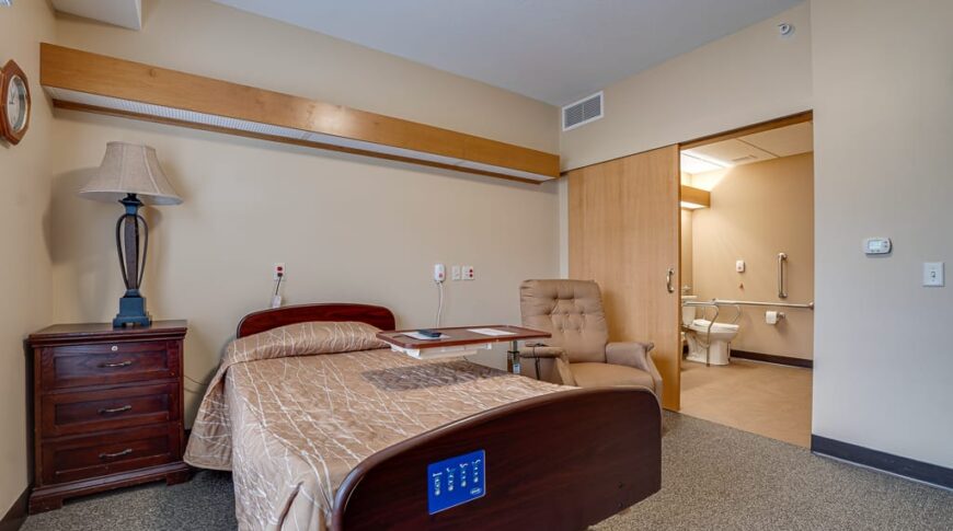 PioneerCare-Low Res-38 Newly remodeled nursing home facility room with medical bed and dresser to one side with rocking chair to other side and sliding door leading to bathroom behind.