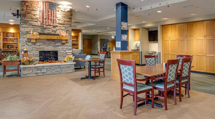 PioneerCare-Low Res-58 New dining room flooring added in dining room at nursing home with tables and chairs throughout backdropped by fireplace behind.