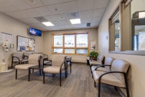 Interior waiting area at Sanford Hillsboro clinic with ample beige-colored chair seating, and three bright windows to the back wall. TV displaying news on back left wall, with bulletin boards underneath.