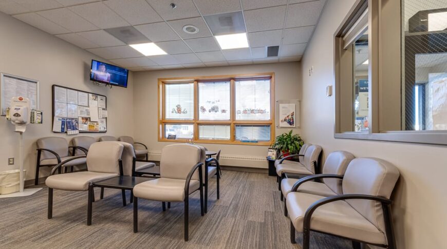 Sanford Hillsboro-Low Res-1 Interior waiting area at Sanford Hillsboro clinic with ample beige-colored chair seating, and three bright windows to the back wall. TV displaying news on back left wall, with bulletin boards underneath.