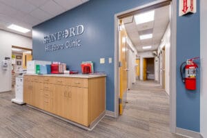 Behind receptionist station at clinic with a blue accent wall with clinic name in metal letters, and light wood-toned cabinetry underneath and medical files. Hallway leading to exam rooms on the back right side.