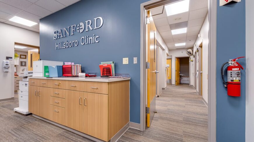 Sanford Hillsboro-Low Res-14 Behind receptionist station at clinic with a blue accent wall with clinic name in metal letters, and light wood-toned cabinetry underneath and medical files. Hallway leading to exam rooms on the back right side.
