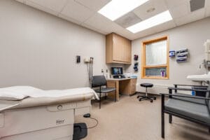 Patient exam room with medical exam table to the left, chairs and sink station to the right and chair and doctor station to the back.