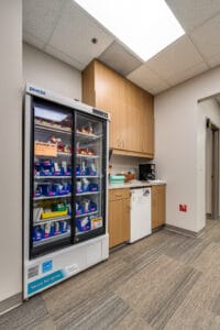 Backroom convenience station for clinic employees with coffee maker and small mini fridge surrounded by upper and lower cabinetry. And a large industrial fridge to the left with refrigerated vaccines.