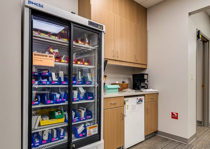 Sanford Hillsboro-Low Res-28 Backroom convenience station for clinic employees with coffee maker and small mini fridge surrounded by upper and lower cabinetry. And a large industrial fridge to the left with refrigerated vaccines.