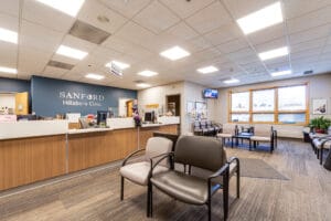 Waiting area at clinic with vinyl-covered chairs in light and dark gray throughout, and a light wood-toned reception station to the back left with a blue-painted accent wall behind with metal words spelling "Sanford Hillsboro Clinic."