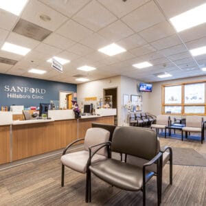 Waiting area at clinic with vinyl-covered chairs in light and dark gray throughout, and a light wood-toned reception station to the back left with a blue-painted accent wall behind with metal words spelling "Sanford Hillsboro Clinic."