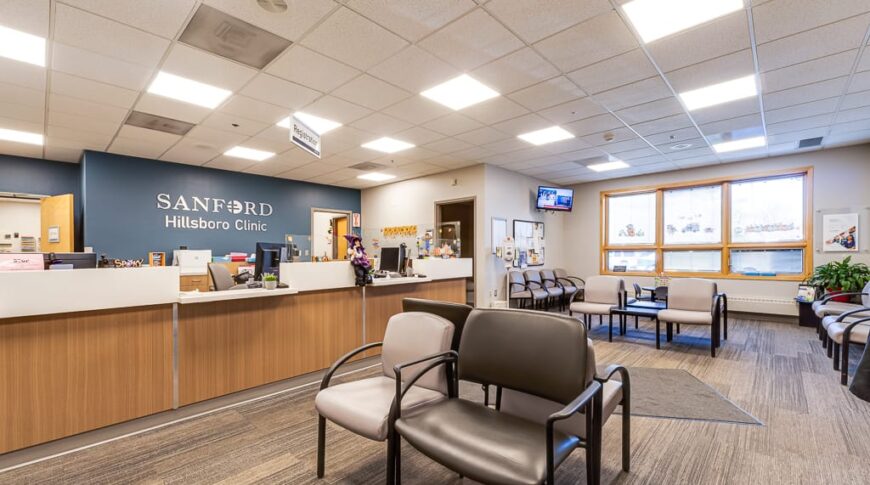 Sanford Hillsboro-Low Res-4 Waiting area at clinic with vinyl-covered chairs in light and dark gray throughout, and a light wood-toned reception station to the back left with a blue-painted accent wall behind with metal words spelling "Sanford Hillsboro Clinic."