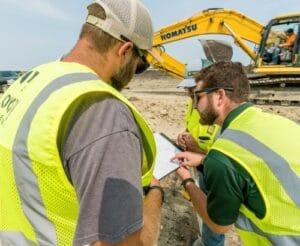 Three men in safety vests pointing at plans while excavator digs in the background.
