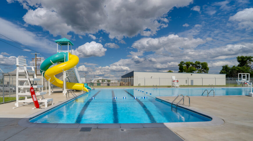 Gwinner Aquatic Center-Low Res-33 Gwinner Aquatic Center swimming pool from deep end viewpoint with lifeguard stand and waterslides to the left and basketball hoop to the right.