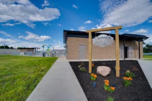 Exterior view of cinder block concession and bathhouse with a triangular-shaped in-ground planter and a metal sign that says "Gwinner Aquatic Center" with a peak of the Splash pad behind.