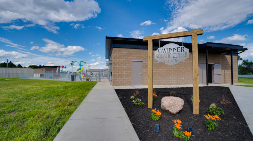 Gwinner Aquatic Center-Low Res-49 Exterior view of cinder block concession and bathhouse with a triangular-shaped in-ground planter and a metal sign that says "Gwinner Aquatic Center" with a peak of the Splash pad behind.