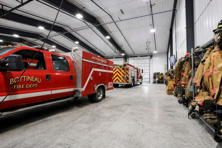 Interior of Bottineau Fire Hall with prefabricated metal building, concrete floors, fire trucks and shelves of firefighters clothing.