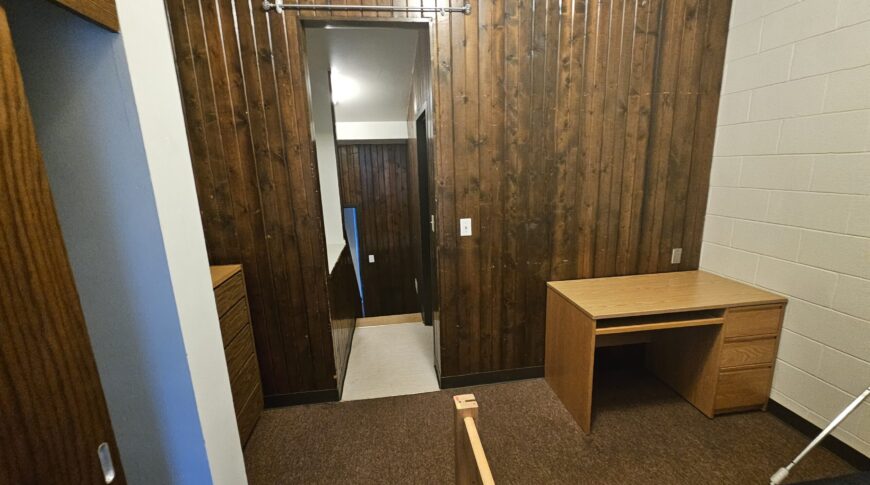 Dorm room at NDSCS College Townhomes with dark wood paneled wall and dark carpeting before renovation.