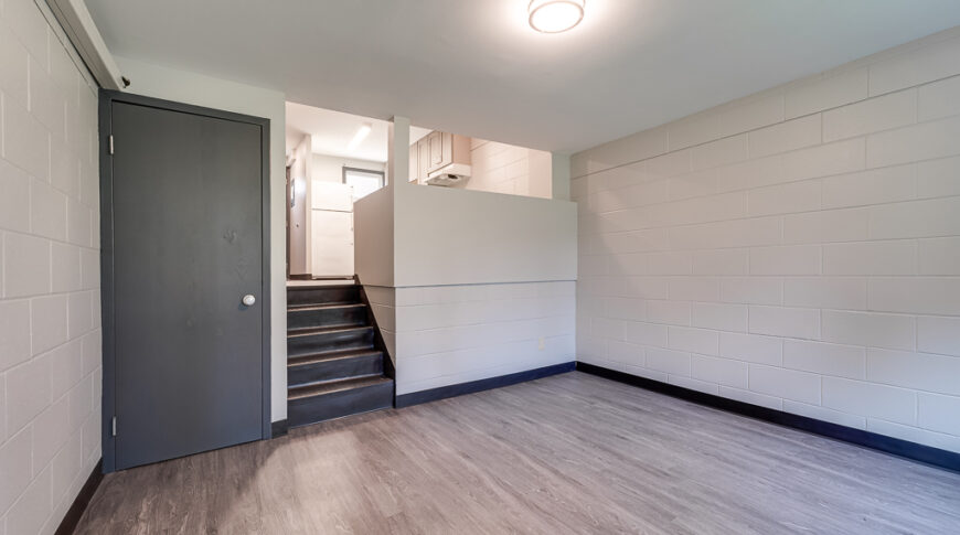 Sunken living room at newly remodeled NDSCS College Townhomes with gray-toned faux-wood LVP Flooring, newly painted white walls, and railing to half-drywall wall into Kitchen.