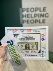 Hand holding up a first-dollar plaque from the Aberdeen Area Chamber of Commerce, with a section of ribbon from the ribbon cutting. Background blurred wall says "Mission: People Helping People."