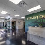 Entryway view of the new Comstock Construction Aberdeen office with dark green vinyl-wrapped wall with the words "Comstock" in gold behind the light-oak reception desk to the right, conference room to the middle of the room, and reception seating area to the left.