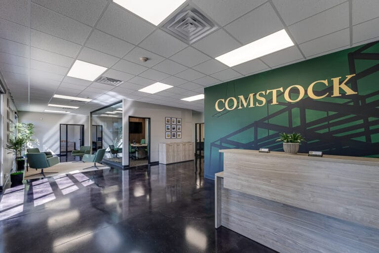 Comstock Construction Aberdeen-Low Res-11 Entryway view of the new Comstock Construction Aberdeen office with dark green vinyl-wrapped wall with the words "Comstock" in gold behind the light-oak reception desk to the right, conference room to the middle of the room, and reception seating area to the left.