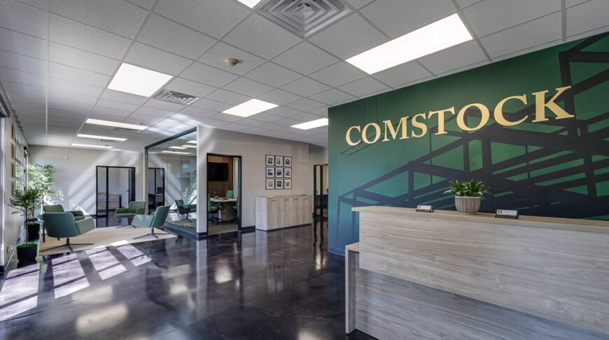 Comstock Construction Aberdeen-Low Res-11 Entryway view of the new Comstock Construction Aberdeen office with dark green vinyl-wrapped wall with the words "Comstock" in gold behind the light-oak reception desk to the right, conference room to the middle of the room, and reception seating area to the left.