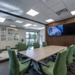 Conference room at Comstock Construction Aberdeen office with light oak table surrounded by green chairs, dark carpeting, wall of windows to the left and wall of acoustic -rated dark walnut wood slats to the right with two large tv monitors mounted.