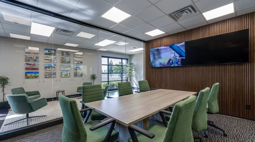 Comstock Construction Aberdeen-Low Res-23 Conference room at Comstock Construction Aberdeen office with light oak table surrounded by green chairs, dark carpeting, wall of windows to the left and wall of acoustic -rated dark walnut wood slats to the right with two large tv monitors mounted.