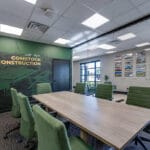 Conference room with large light-oak table in the middle surrounded by green chairs, with wall of floor-to-ceiling windows behind overlooking reception area and a branded vinyl-wrapped dark green accent wall with the words "We are Comstock Construction" to the back.