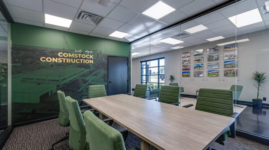 Comstock Construction Aberdeen-Low Res-24 Conference room with large light-oak table in the middle surrounded by green chairs, with wall of floor-to-ceiling windows behind overlooking reception area and a branded vinyl-wrapped dark green accent wall with the words "We are Comstock Construction" to the back.