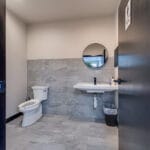Single private bathroom space with toilet and sink and mirror with greige walls and gray and white large-format tiles on the floor and the backsplash.