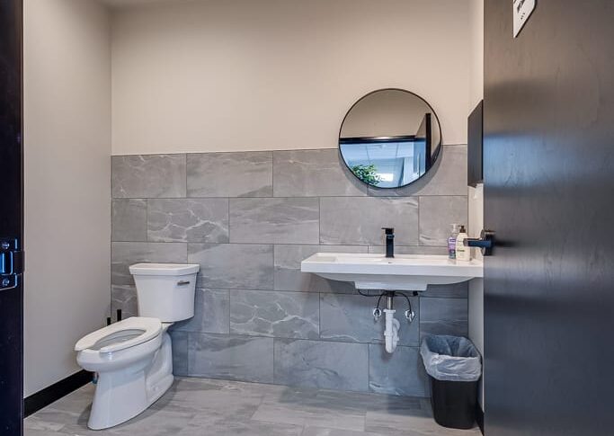 Comstock Construction Aberdeen-Low Res-35 Single private bathroom space with toilet and sink and mirror with greige walls and gray and white large-format tiles on the floor and the backsplash.