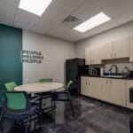 Breakroom at Comstock Construction Aberdeen office with light oak cabinetry and black countertops to the right, a circular dining tables surrounded by green chairs, and an accent wall behind with a green stripe down the wall with the words "Mission: People Helping People."