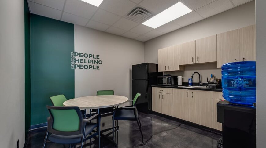 Comstock Construction Aberdeen-Low Res-37 Breakroom at Comstock Construction Aberdeen office with light oak cabinetry and black countertops to the right, a circular dining tables surrounded by green chairs, and an accent wall behind with a green stripe down the wall with the words "Mission: People Helping People."