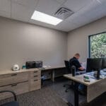 Office with greige walls, light wood-toned cabinetry (only lowers), and a window overlooking green bushes.
