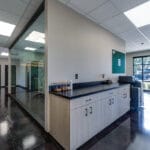 Corner intersection of office with business station cabinets and printer to the right and wall of windows overlooking the conference room to the left.