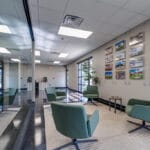 Office reception space with four modern green chairs with jute rug underneath, hanging cable gallery wall of past project pictures besides, and floor to ceiling wall of windows overlooking conference room to the other side.