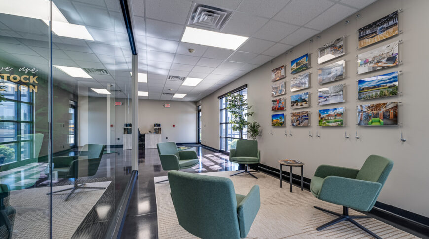 Comstock Construction Aberdeen-Low Res-61 Office reception space with four modern green chairs with jute rug underneath, hanging cable gallery wall of past project pictures besides, and floor to ceiling wall of windows overlooking conference room to the other side.