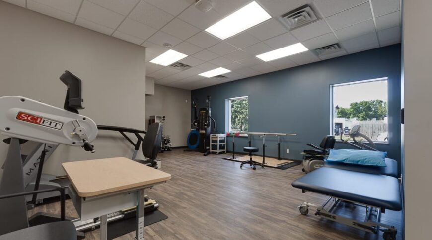 Sanford Hankinson-Low Res-17 Physical therapy room with dark blue accented back wall and light greige walls besides, with medium wood-colored LVP flooring throughout as well as physical therapy equipment throughout.