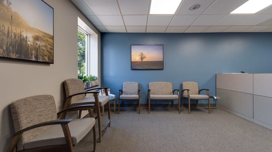 Sanford Hankinson-Low Res-3 Seating waiting area at clinic with blue-grey back wall and greige colored left wall. A couple landscaped framed pictures for accents, and some greenery arrangements in the back left window.