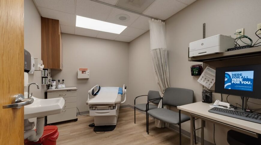 Sanford Hankinson-Low Res-39 Exam room at Sanford Hankinson clinic with exam table to the back with privacy curtain and two chairs besides. Sink wash station, cabinetry, and small desktop station with computer also in the room.