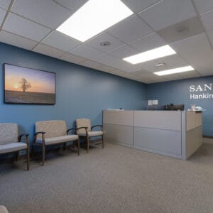Entry reception area at Sanford Hankinson Clinic with blue-grey colored walls, scenic picture hung on back left wall, waiting area chairs, and reception desk station to the back left wall with the words "Sanford Hankinson Clinic" in metal letters on the wall.