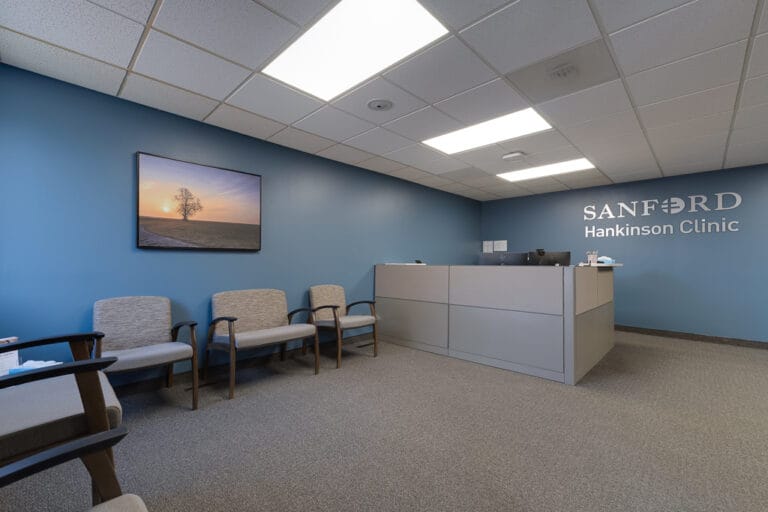Sanford Hankinson-Low Res-5 Entry reception area at Sanford Hankinson Clinic with blue-grey colored walls, scenic picture hung on back left wall, waiting area chairs, and reception desk station to the back left wall with the words "Sanford Hankinson Clinic" in metal letters on the wall.