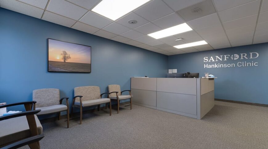 Sanford Hankinson-Low Res-5 Entry reception area at Sanford Hankinson Clinic with blue-grey colored walls, scenic picture hung on back left wall, waiting area chairs, and reception desk station to the back left wall with the words "Sanford Hankinson Clinic" in metal letters on the wall.