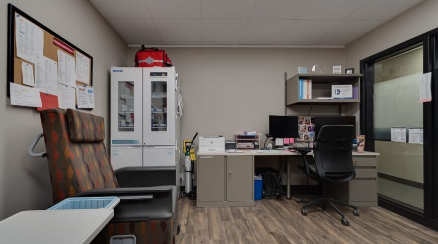 Sanford Hankinson-Low Res-50 Lab draw station with desk and rolling chair to the back right wall, cabinetry with medical supplies to back left wall, and chair for lab draws in foreground.